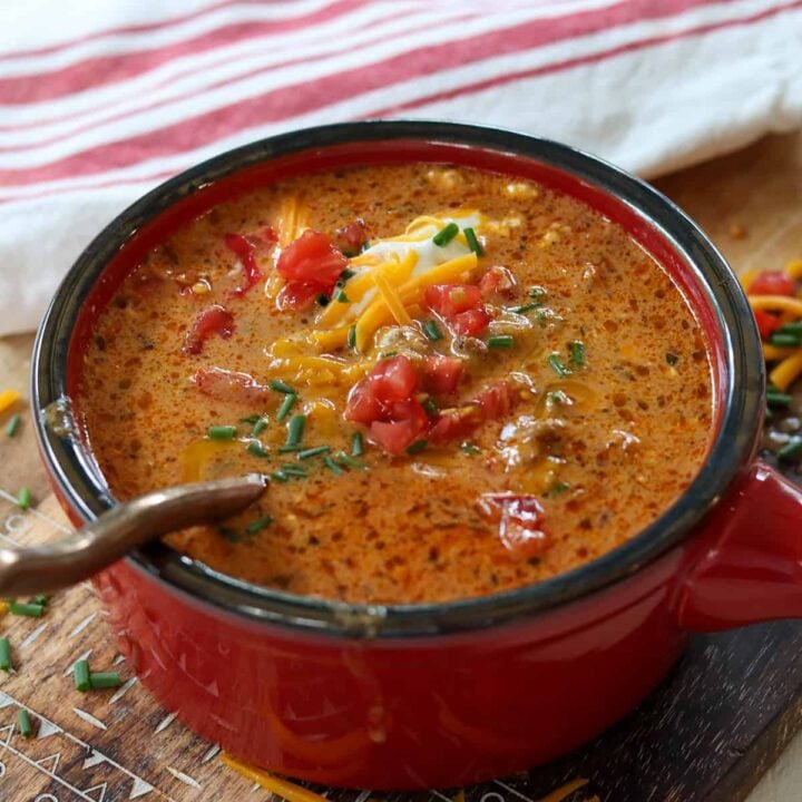 Image côté carré de soupe servie dans un bol rouge sur une planche à découper en bois. La soupe est garnie de crème sure, de tomate, d'oignon vert et de fromage cheddar. Une cuillère est prête pour que vous preniez une bouchée.