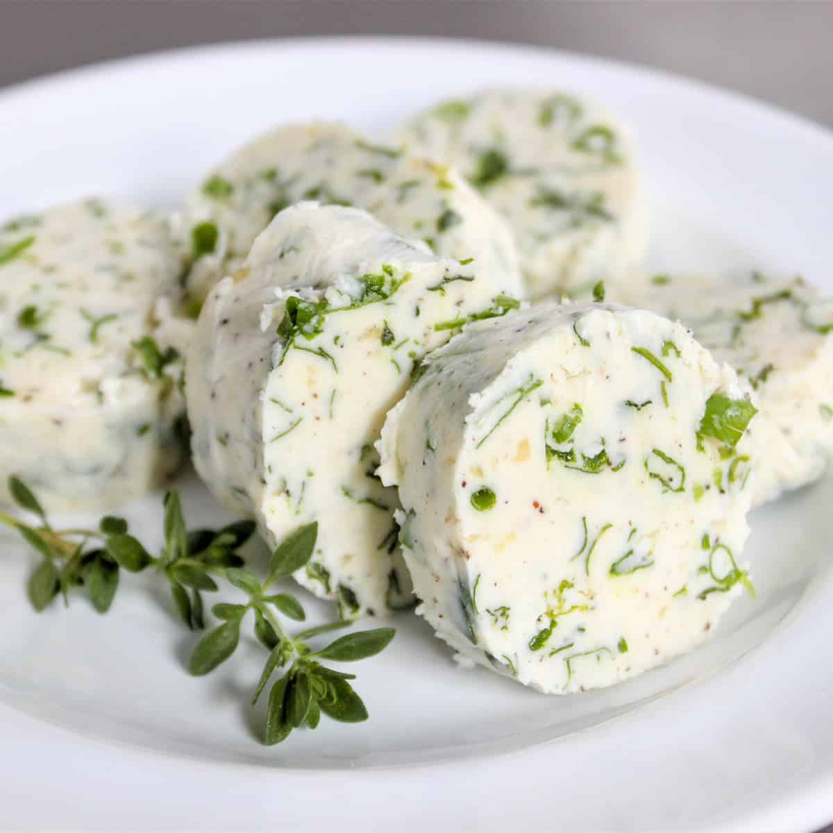Square closeup image of garlic herb compound butter served on a white plate. It is sliced into pats of butter and garnished with thyme