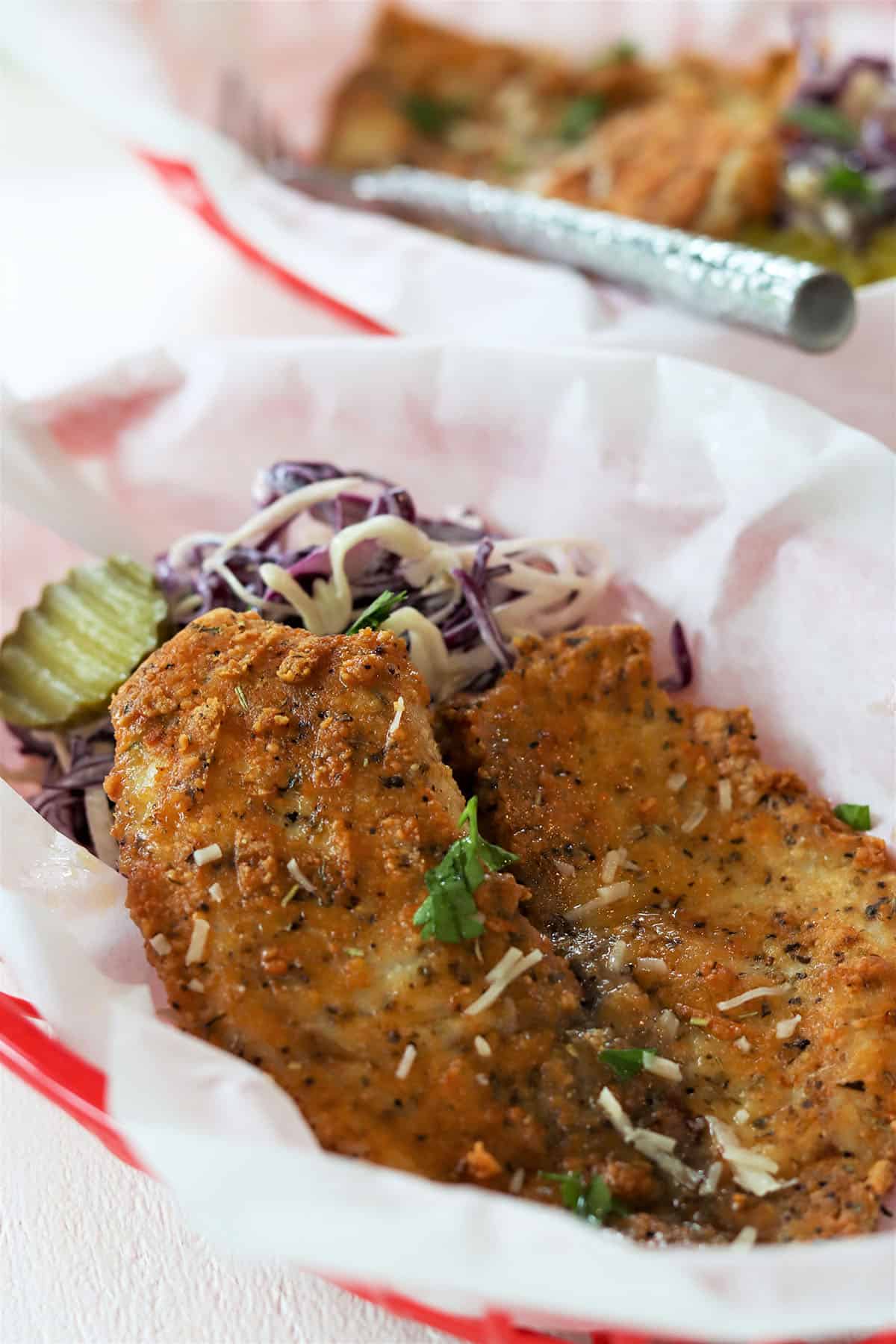 A golden piece of parmesan crusted tilapia in a red basket lined with white parchment paper with a side of coleslaw and pickles.