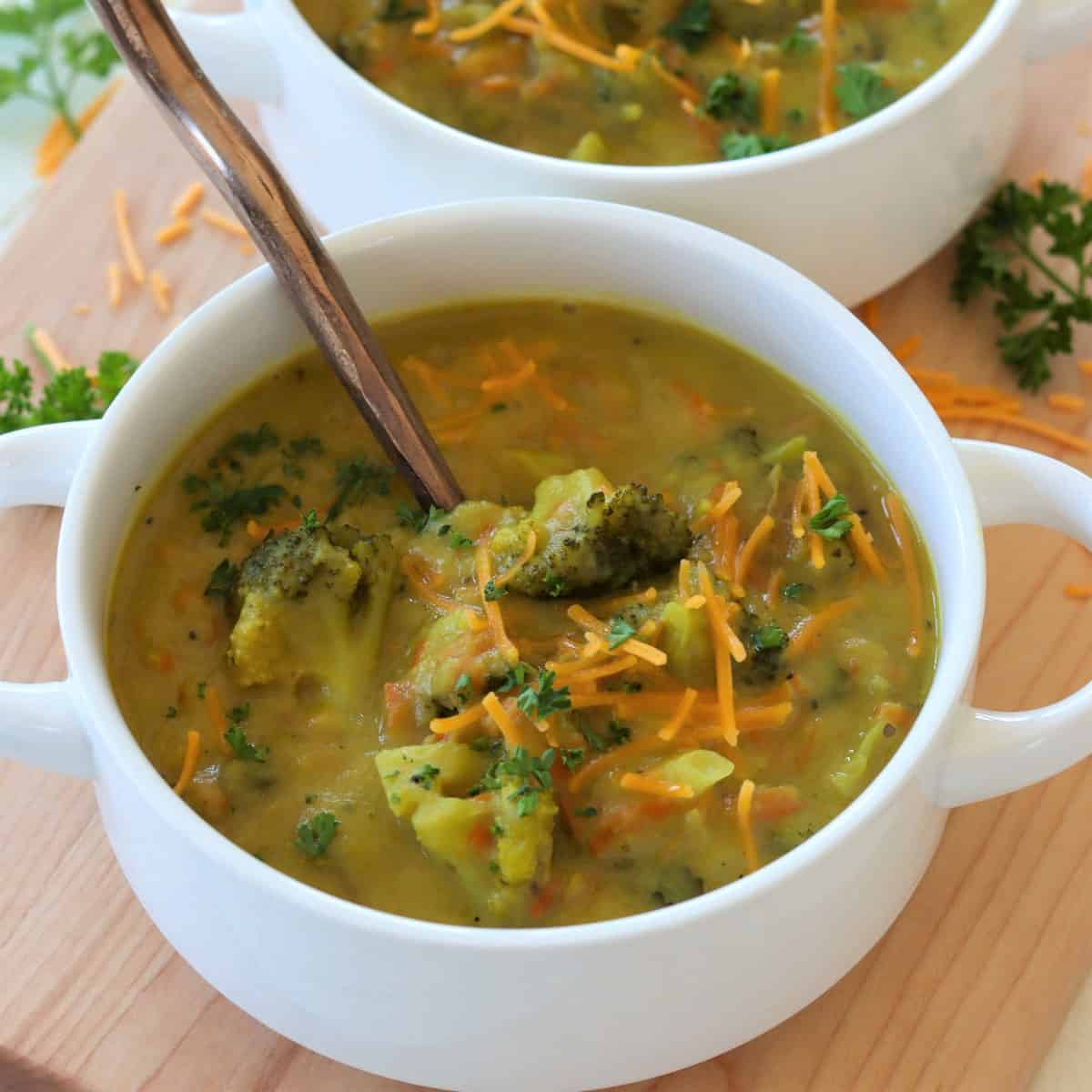 Vegan broccoli soup served in a white bowl with handles and garnished with vegan cheddar cheese.