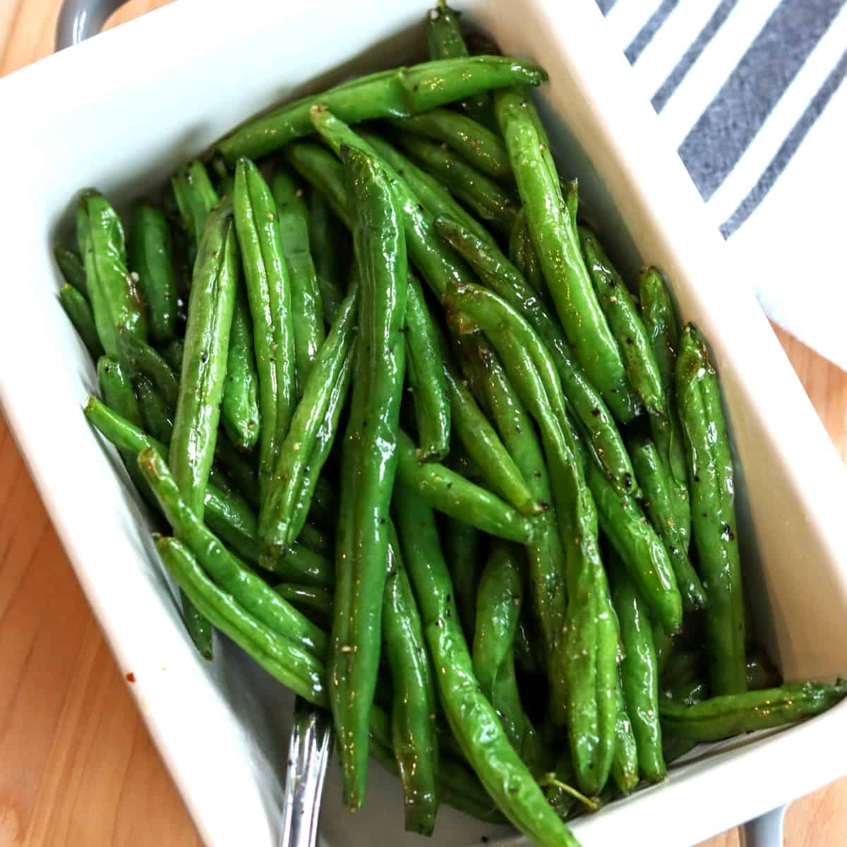 green beans served in a whtie oval dish with a spoon and a blue and white napkin on the side.
