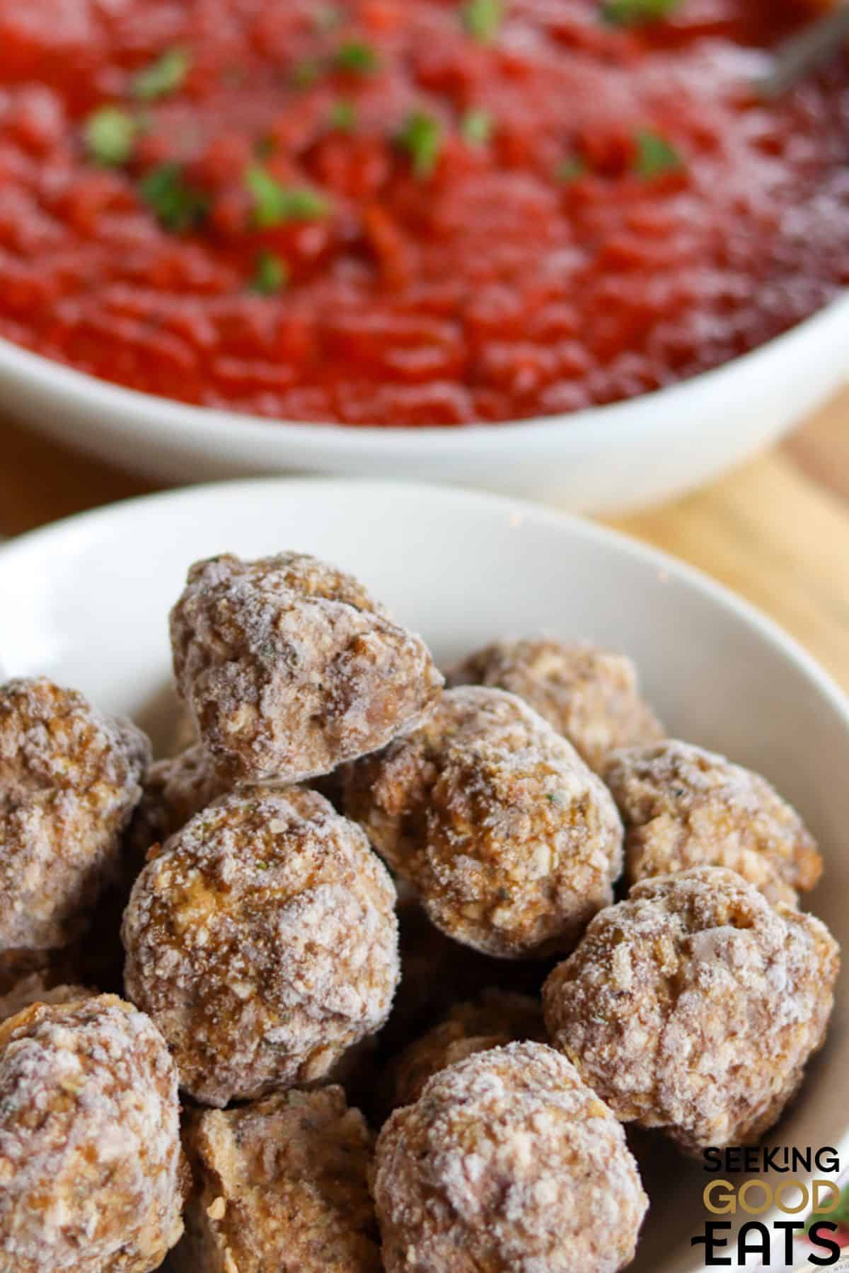 Homemade meatballs in a bowl next to a bowl of marinara sauce.