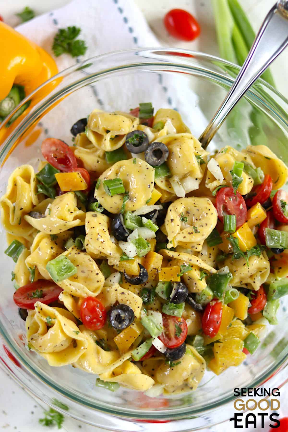 A bowl of creamy tortellini pasta salad with fresh tomatoes and black olives being served with a spoon.