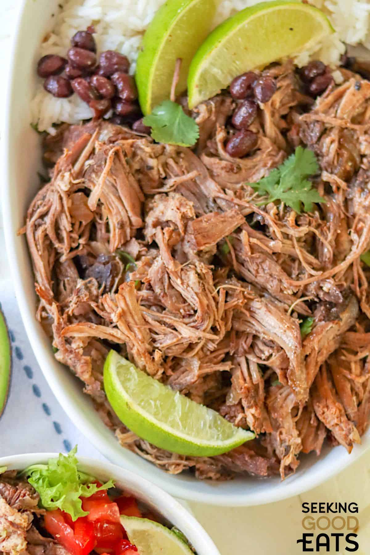 Tender shredded barbacoa with lime wedges and beans in a white bowl.