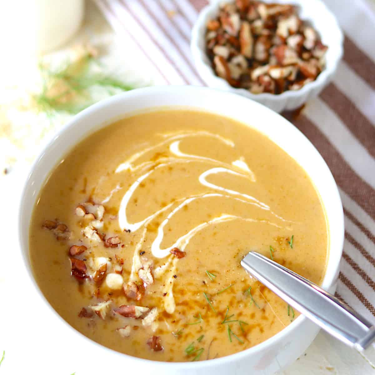 A white bowl of butternut squash soup drizzled with sour cream and topped with toasted pecans.