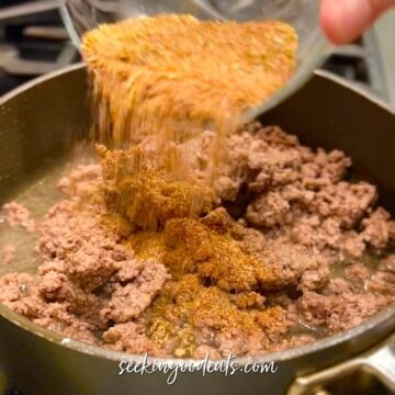 Adding taco seasoning mix to cooked ground beef in a dark skillet.