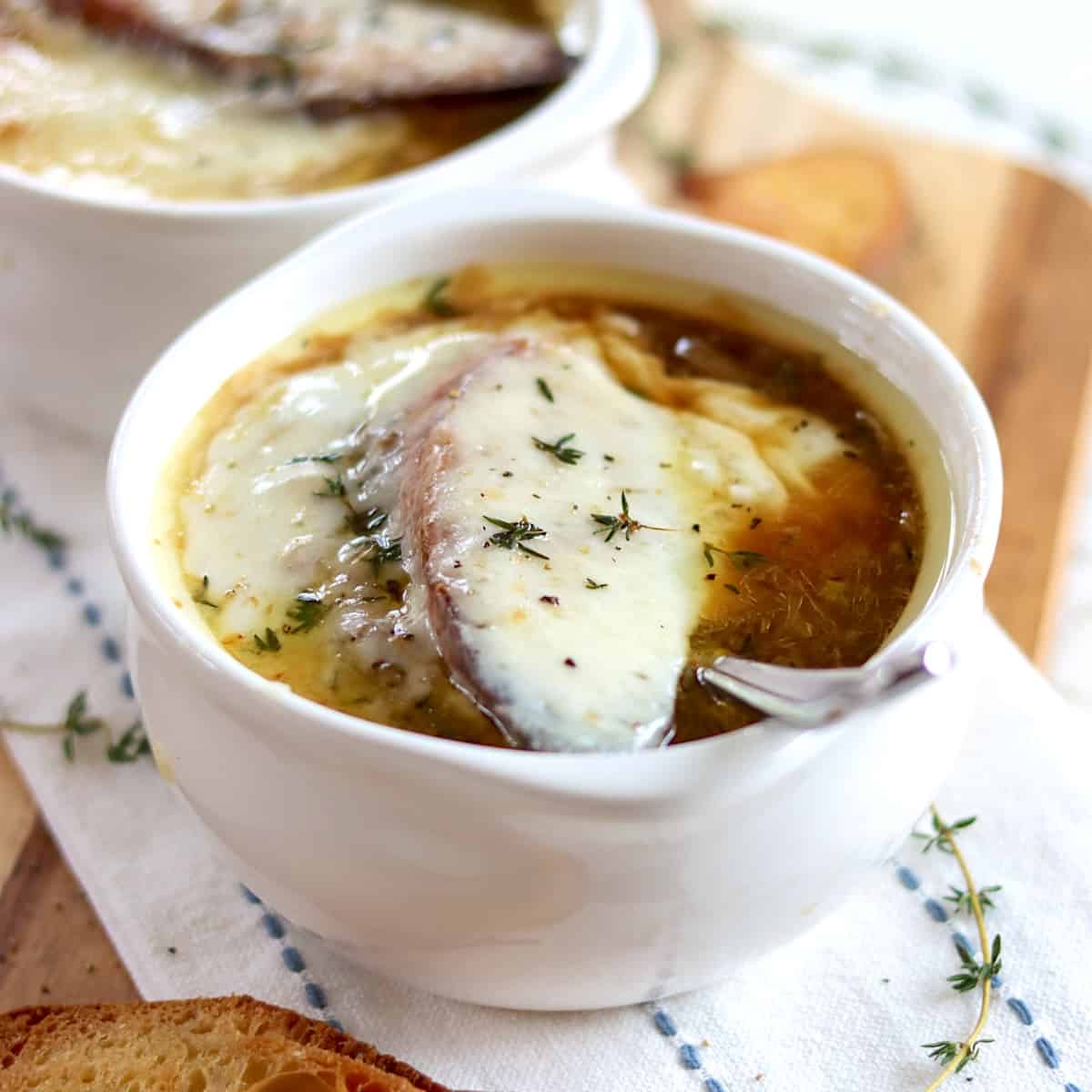 Gooey melted cheese over a crouton floating on French onion soup in a white crockery bowl.