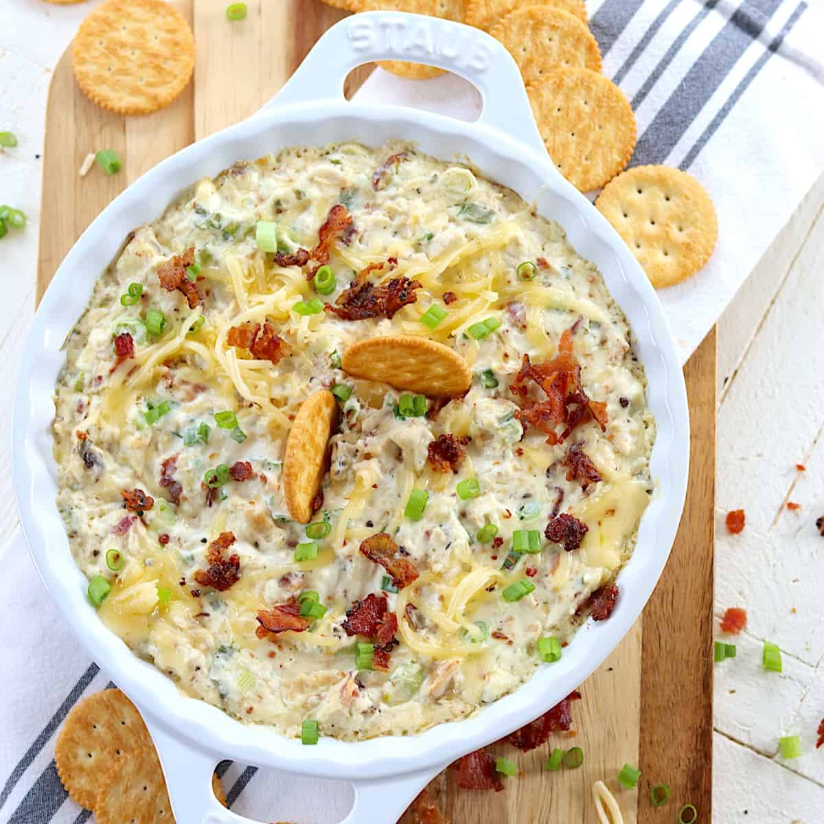 A white handled baking dish with hot cheesy crack chicken dip topped with bacon and green onion.