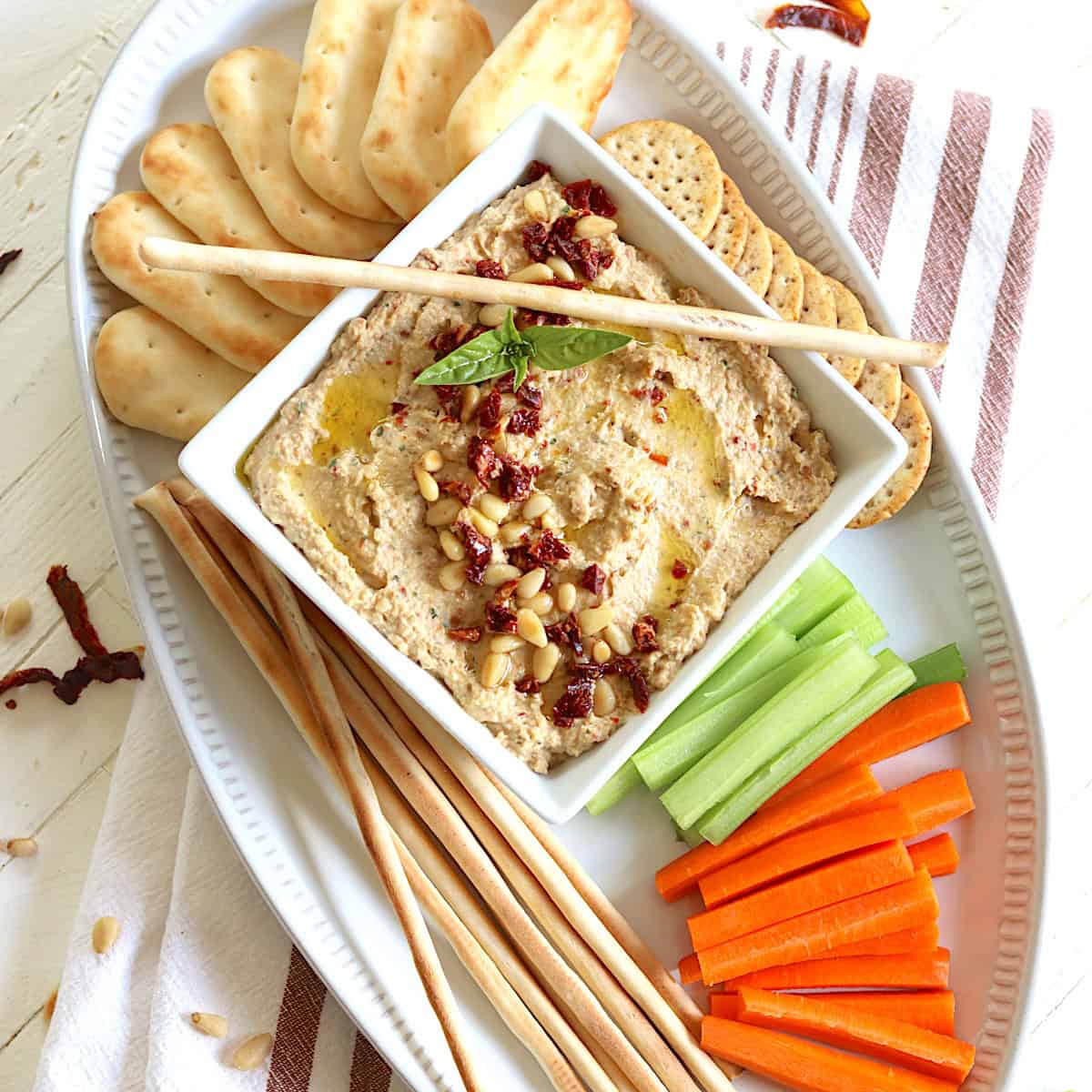 A white oval platter with crackers and raw veggies for dipping in a bowl of hummus dip.