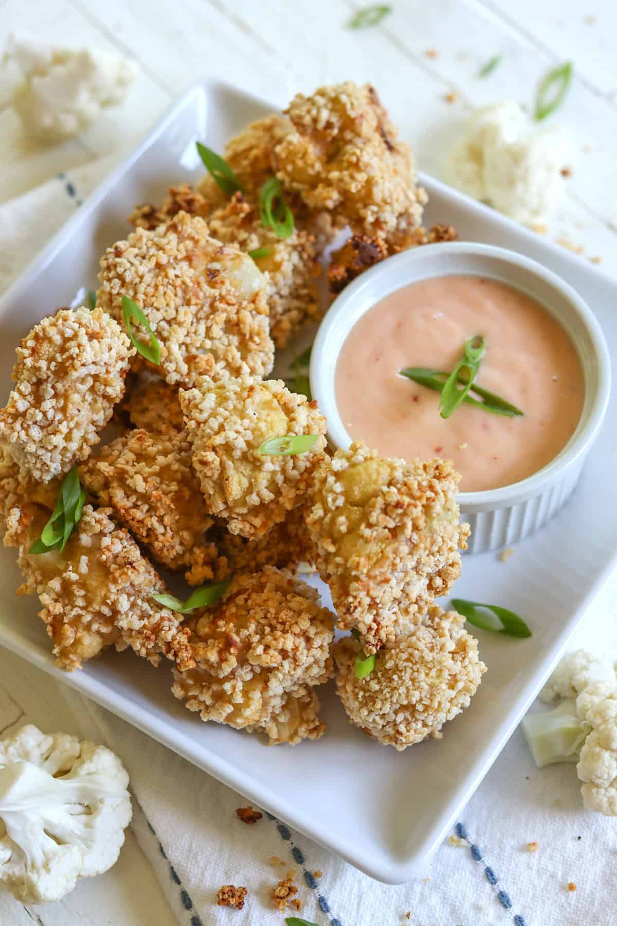 A square white plate piled high with crispy bang bang cauliflower with dipping sauce.