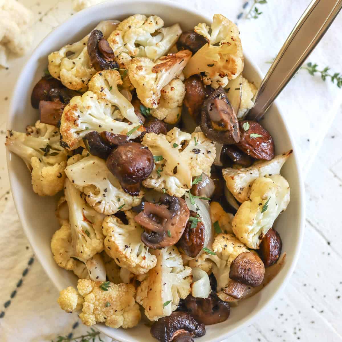 Golden roasted cauliflower and mushrooms with onions and herbs in a white bowl with a spoon.