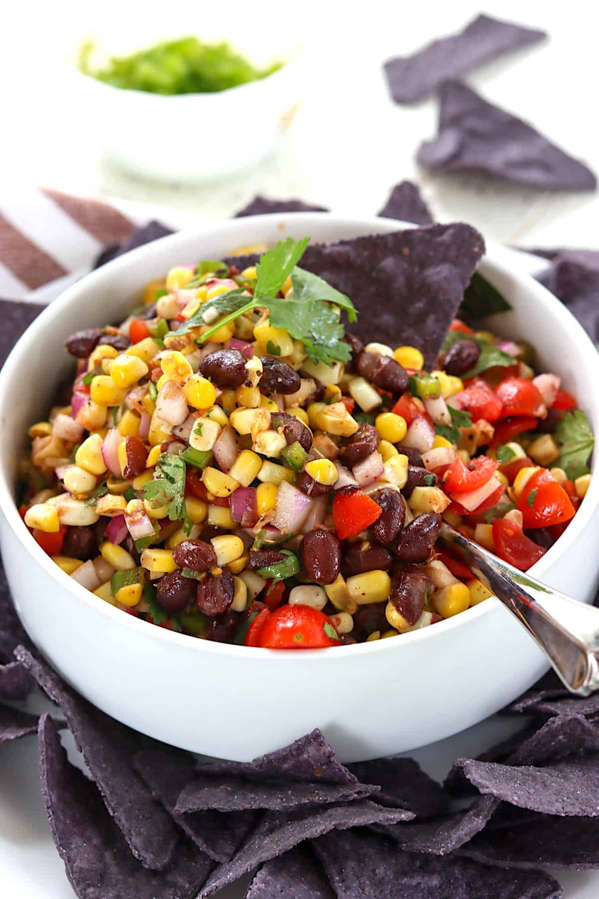 A white serving bowl with black and bean and corn salsa with a spoon and tortilla chips.