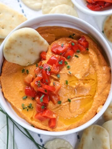 Homemade smoky red bell pepper hummus in a white bowl with a cracker for dipping.