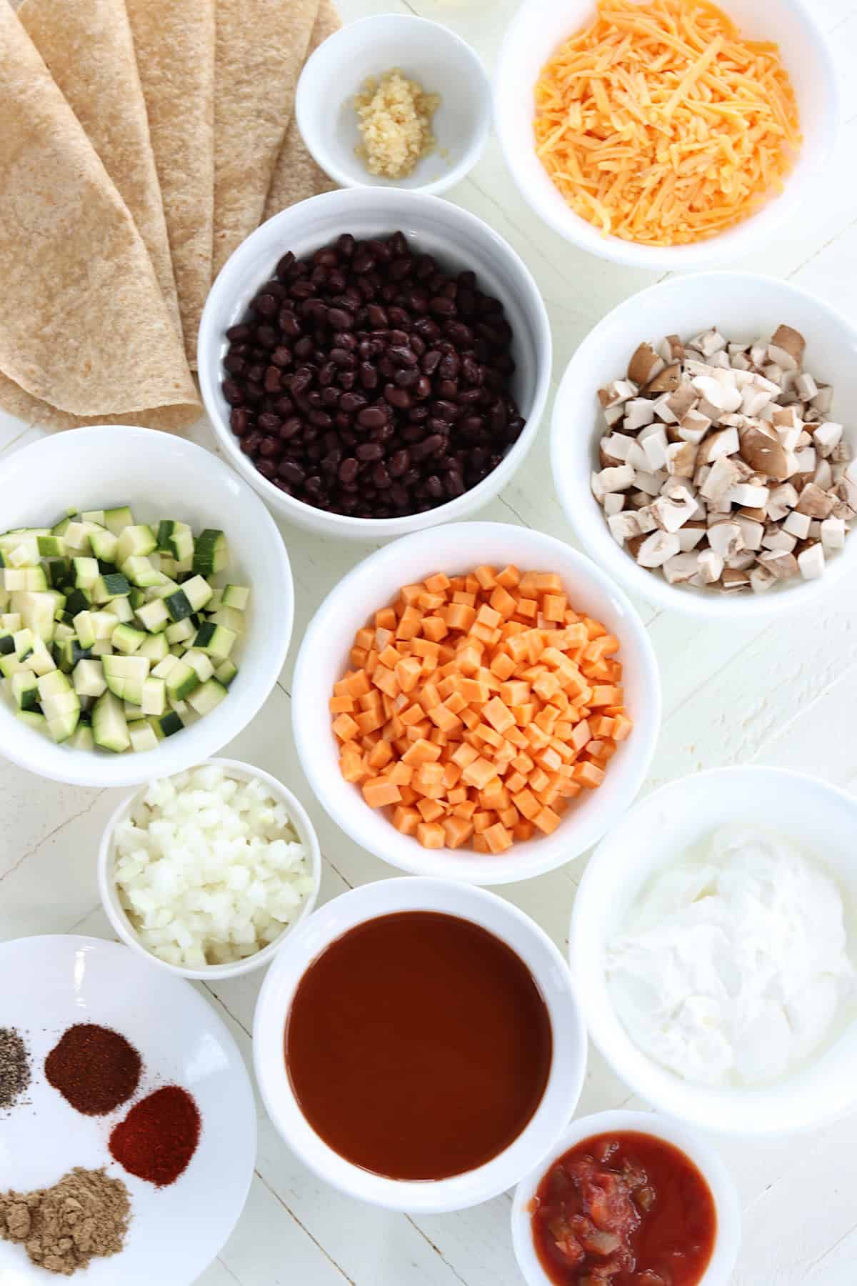 Ingredients need for making black bean sweet potato enchiladas on a white wooded board.