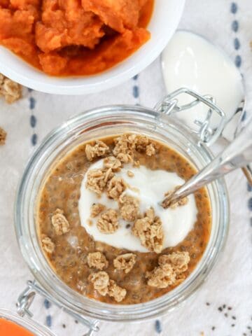 A glass lidded jar of pumpkin chia pudding with a spoon and a granola topping.