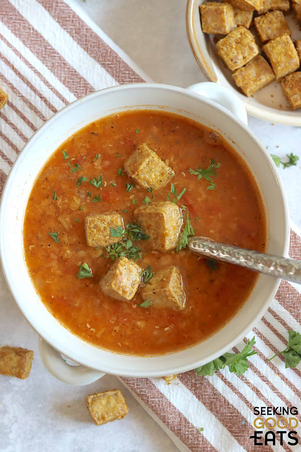 Red lentil and tomato soup topped with tofu croutons and fresh parsley with a spoon.