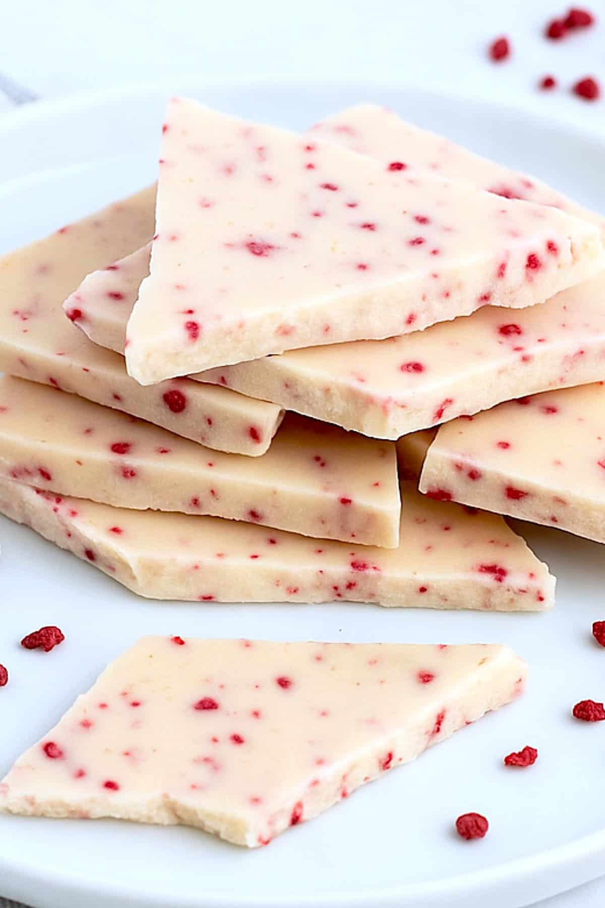 Raspberry white chocolate bark pieces stacked on a plate with scattered raspberries.