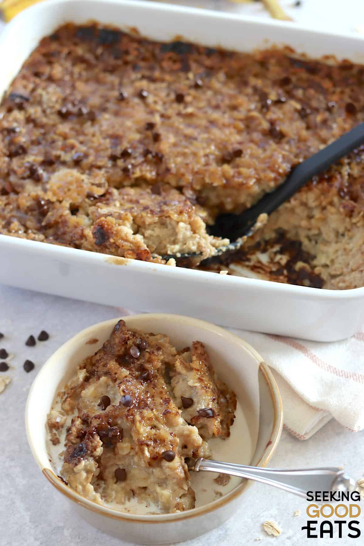 Banana chocolate chip baked oatmeal in a cream bowl and silver spoon next to a white rectangle serving dish.
