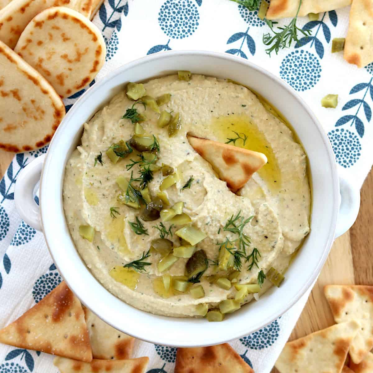 Easy dill pickle hummus served in a white round crock and garnished with diced dill pickles and served with pita chips.