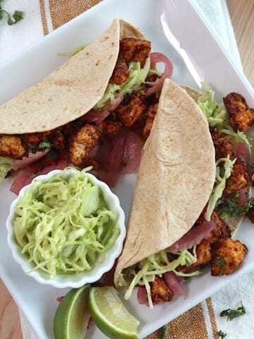 Two tofu tacos on a white square plate with two lime wedges and a small bowl of slaw on the plate.