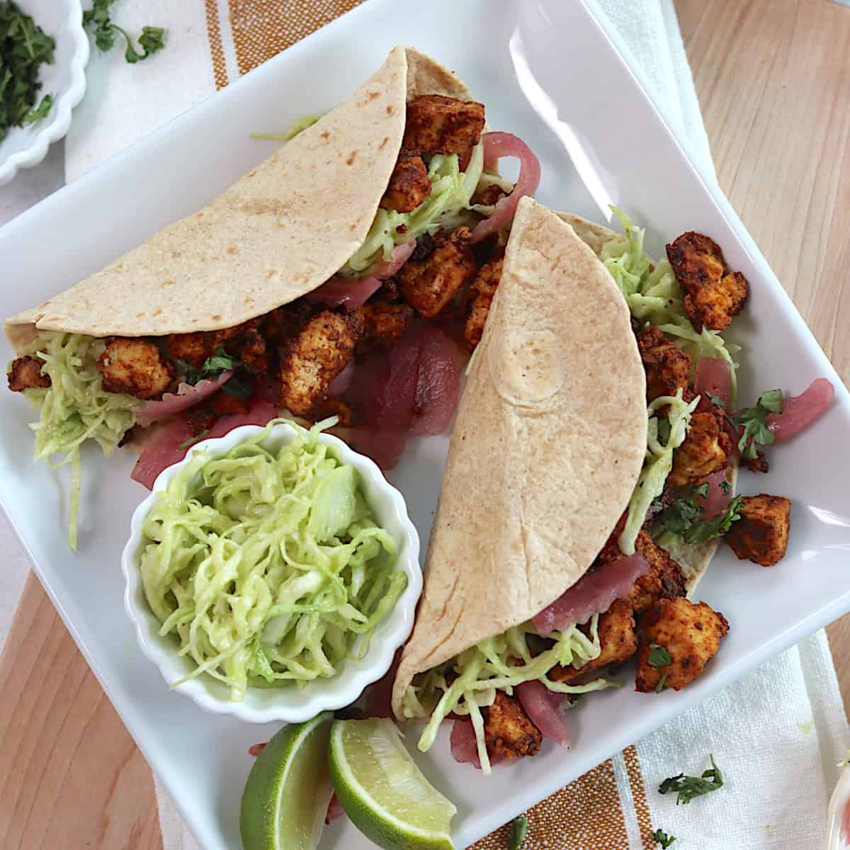 Two tofu tacos on a white square plate with two lime wedges and a small bowl of slaw on the plate.