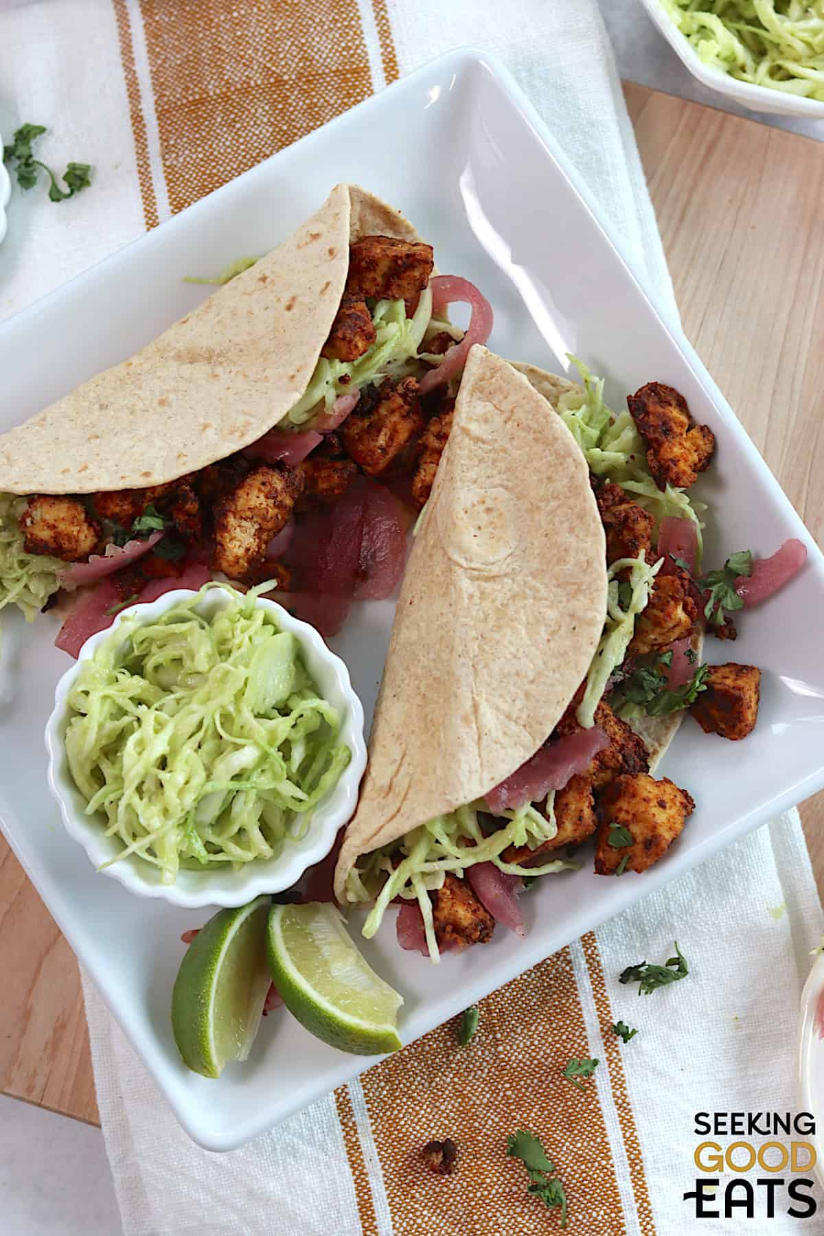 Two tofu tacos on a white square plate with two lime wedges, and a small white bowl of slaw.