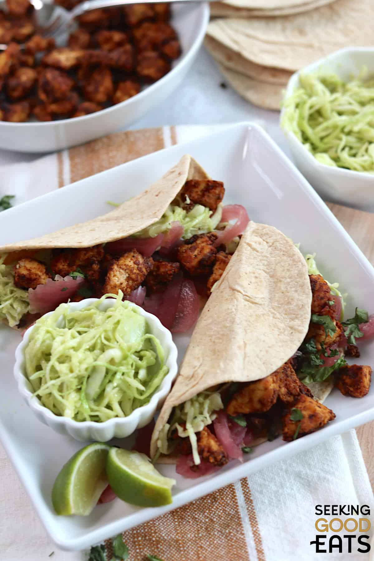 Two tofu tacos on a white square plate with a small white bowl of slaw, and two lime wedges.