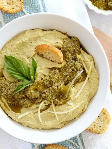 Creamy pesto hummus in a white round bowl, topped with basil pesto.