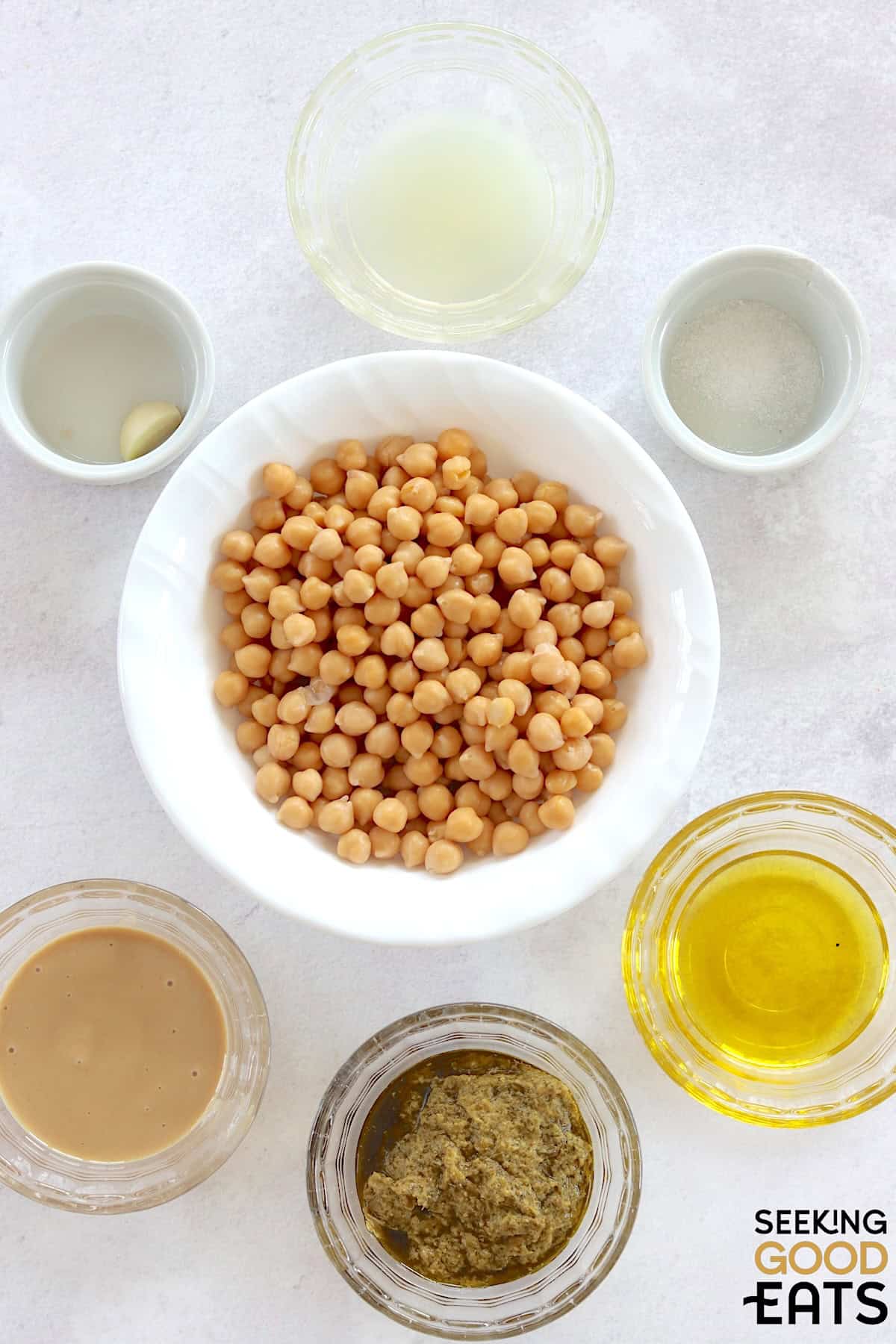Ingredients needed to make pesto hummus arranged on a white marble countertop.