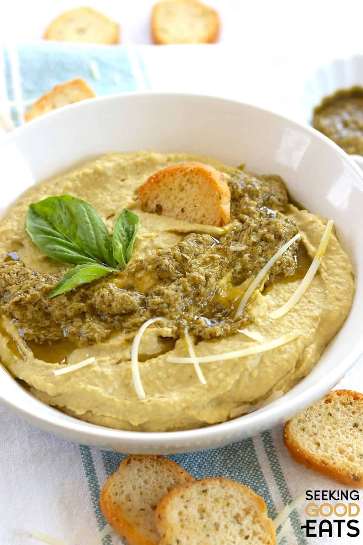 Creamy pesto hummus in a white round bowl, topped with basil pesto.