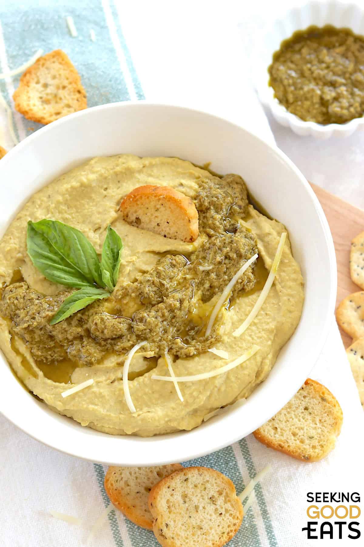 Creamy pesto hummus in a white round bowl, topped with basil pesto.