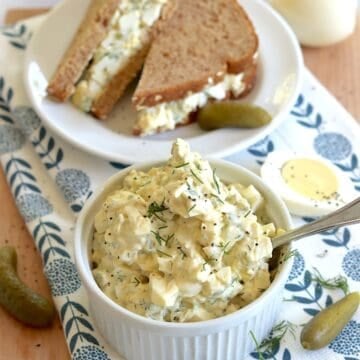 Egg salad with dill pickles served in a small white ramekin and a silver spoon, next to an egg salad sandwich on a small round white plate.
