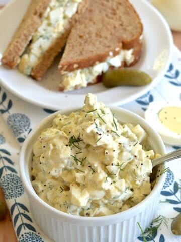 Egg salad with dill pickles served in a small white ramekin and a silver spoon, next to an egg salad sandwich on a small round white plate.