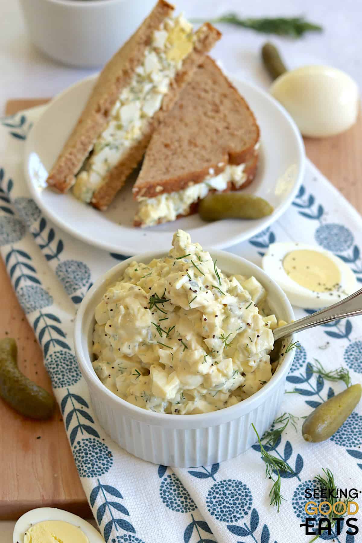 Egg salad with dill pickles in small white ramekin and a silver spoon, next to an egg salad sandwich on a small round white plate.