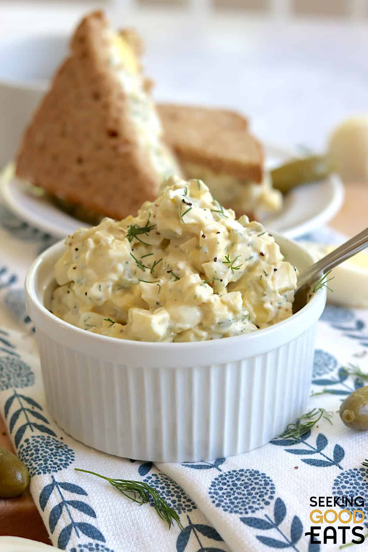 Egg salad with dill pickles in a small white ramekin and a silver spoon.