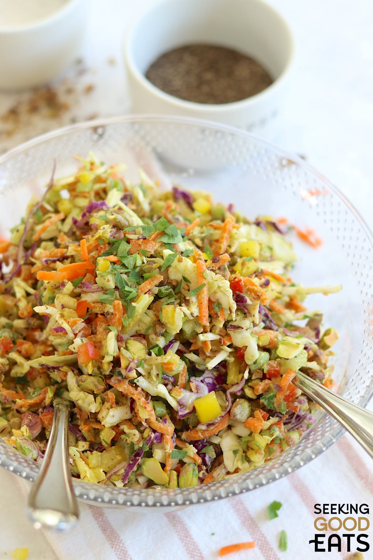A large round clear bowl filled with green goddess cabbage salad and two silver salad spoons.