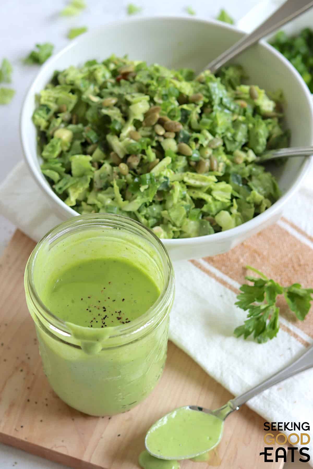 Green goddess dressing recipe served in a clear mason jar and a sprinkle of black pepper, next to a white bowl filled with green goddess salad.