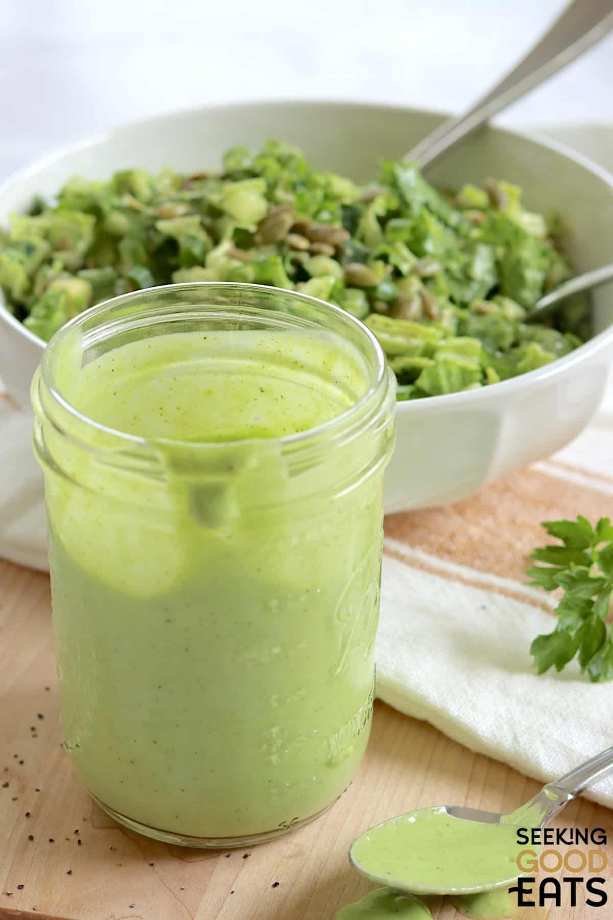 A clear mason jar filled with green goddess dressing recipe, next to a white bowl filled with green goddess salad.