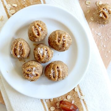 Six date protein balls served on a white round plate, drizzled with peanut butter and topped with flaky salt.