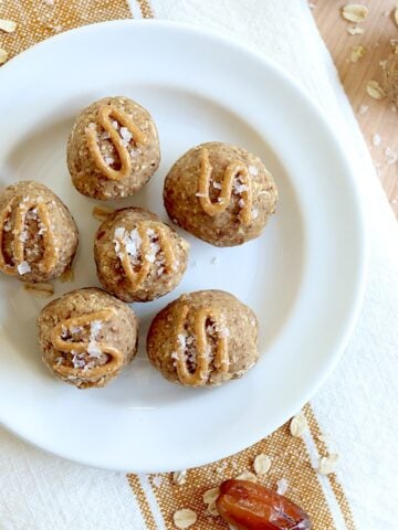 Six date protein balls served on a white round plate, drizzled with peanut butter and topped with flaky salt.