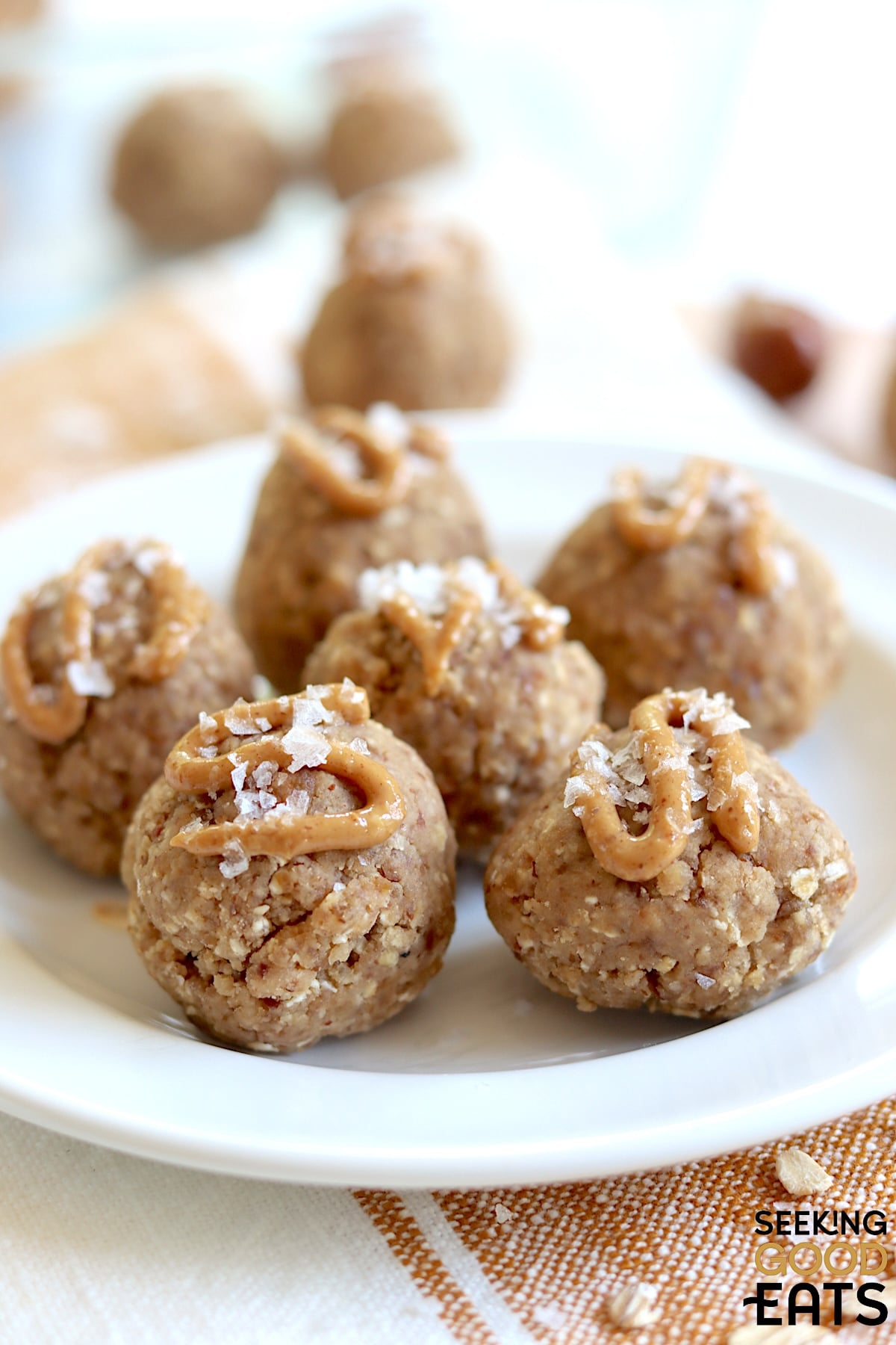 A close up of date protein balls on a round white plate, drizzled with peanut butter and topped with flaky salt.