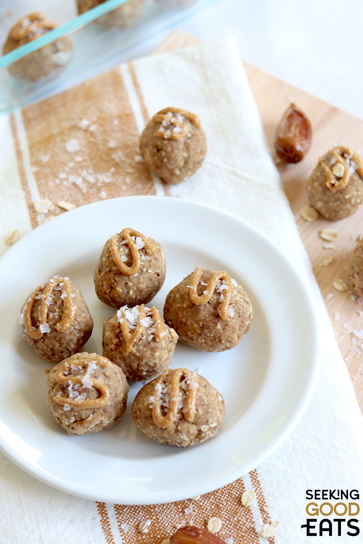 Six date protein balls arranged on a round white plate drizzled with peanut butter and topped with flaky salt.