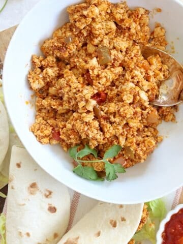 Plant based tofu taco meat in a round white bowl and a silver spoon next to two soft shell tacos.