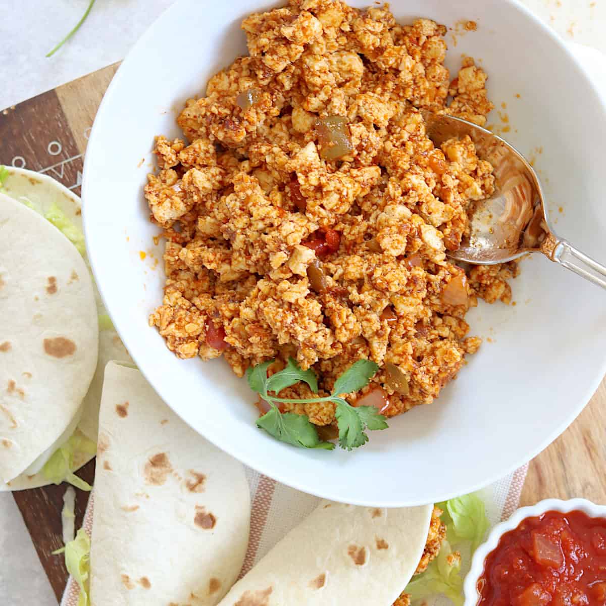 Plant based tofu taco meat in a round white bowl and a silver spoon next to two soft shell tacos.