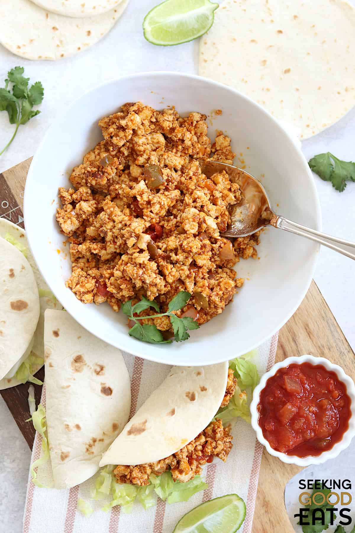 Plant based tofu taco meat in a round white bowl and a silver spoon, with two soft shell tacos on the side.