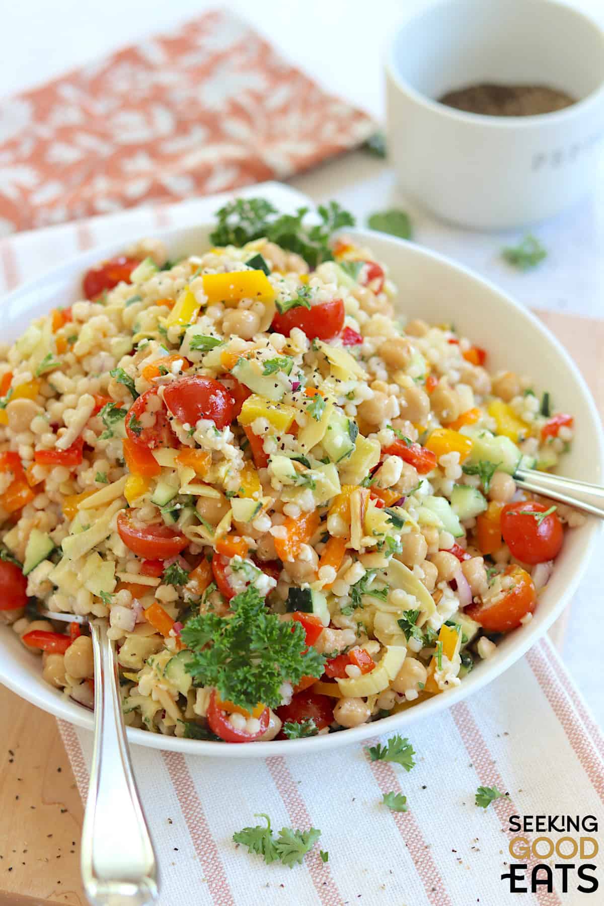 Mediterranean pearl couscous salad served in a shallow round white bowl and two silver serving spoons.