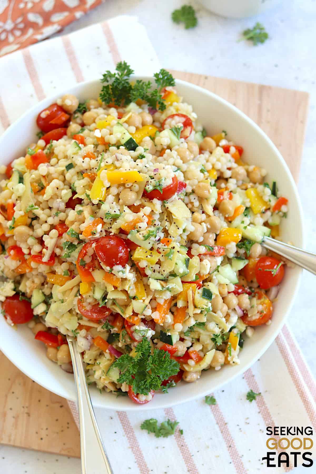 Mediterranean pearl couscous salad served in a shallow round white bowl and two silver serving spoons.