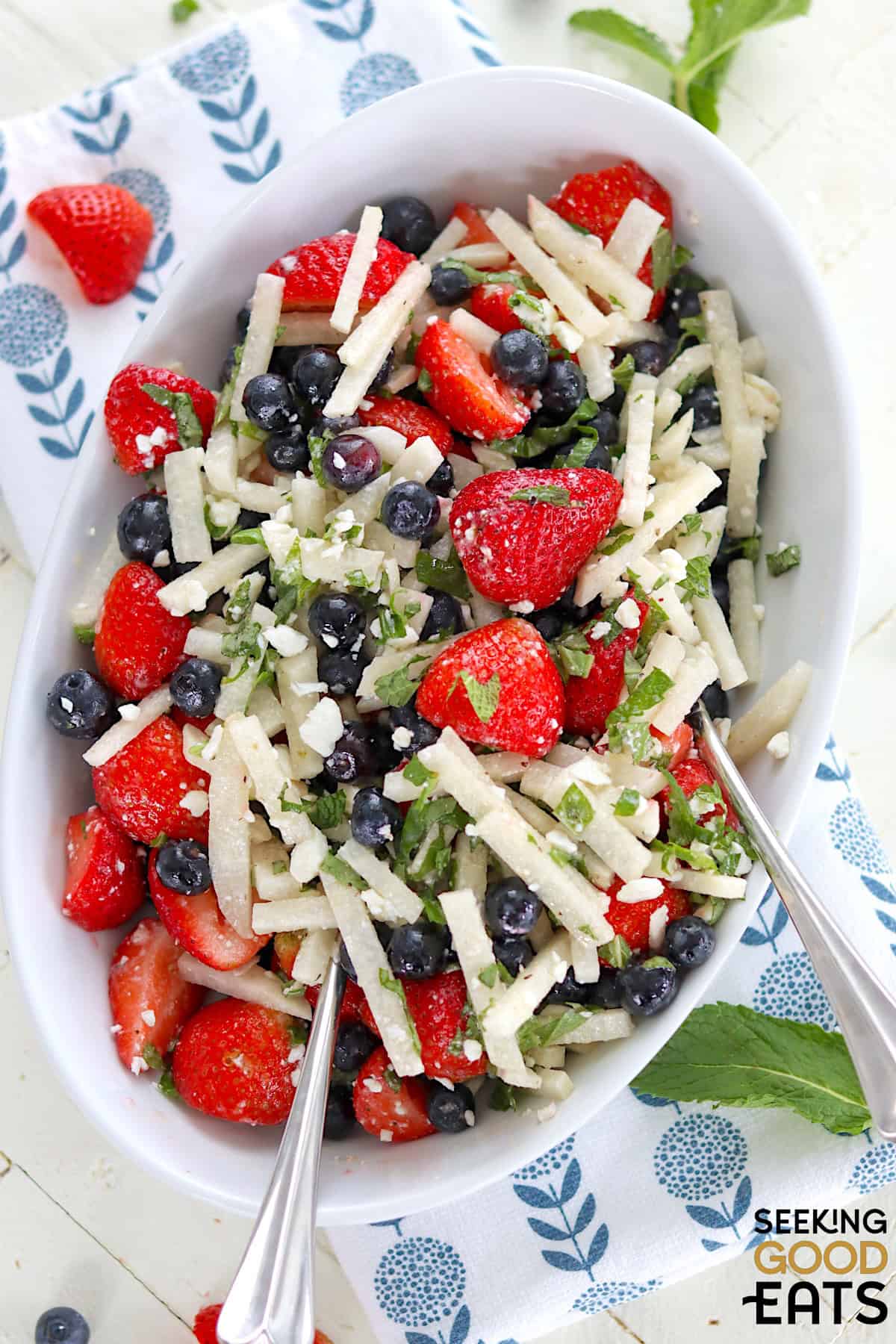 Serving the red white and blue fruit salad in a white oval serving bowl and two silver serving spoons.