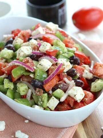Chunky fresh Greek salad in a white oval bowl drizzled with dressing.