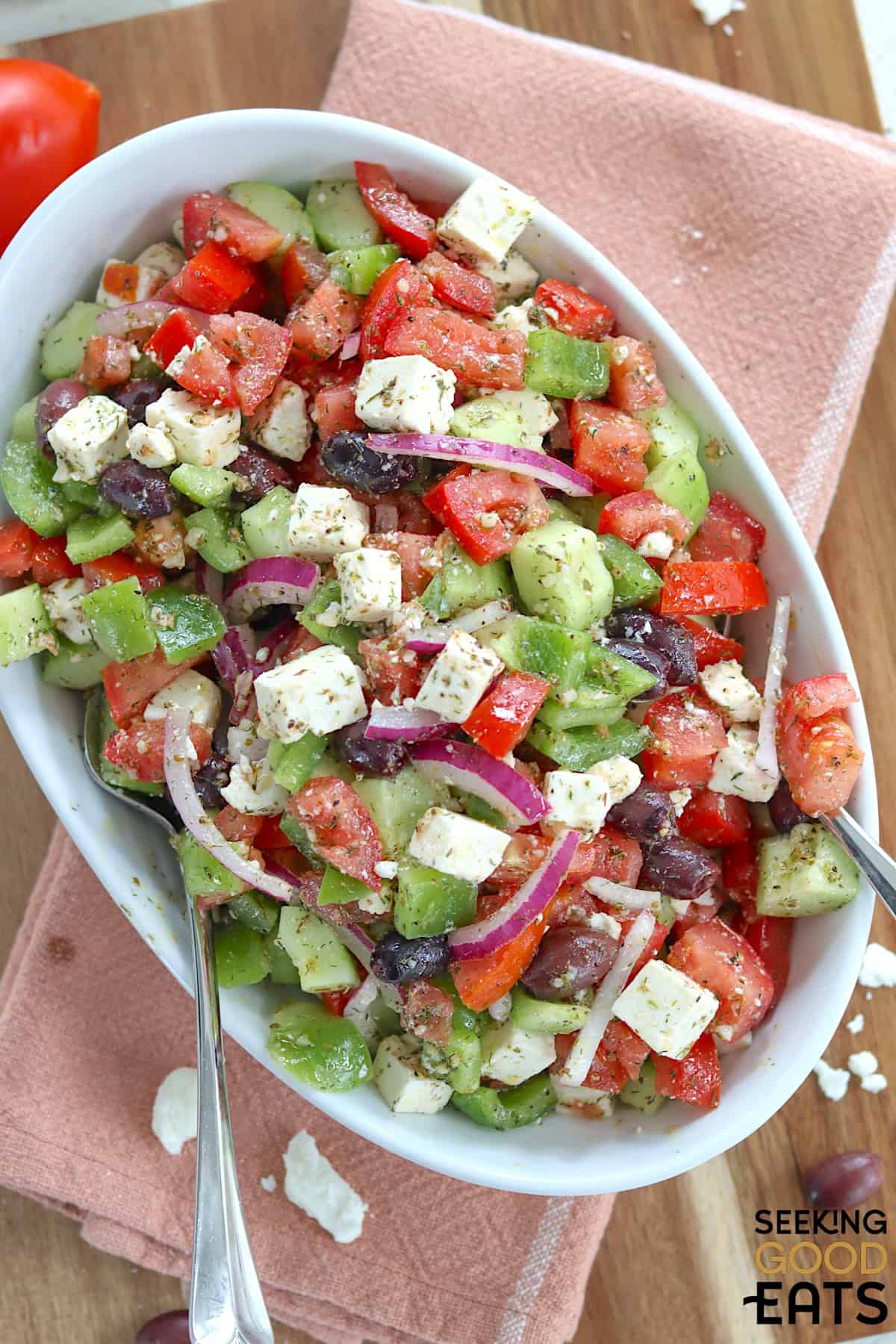 An oval white salad bowl with traditional Greek salad and serving utensils.